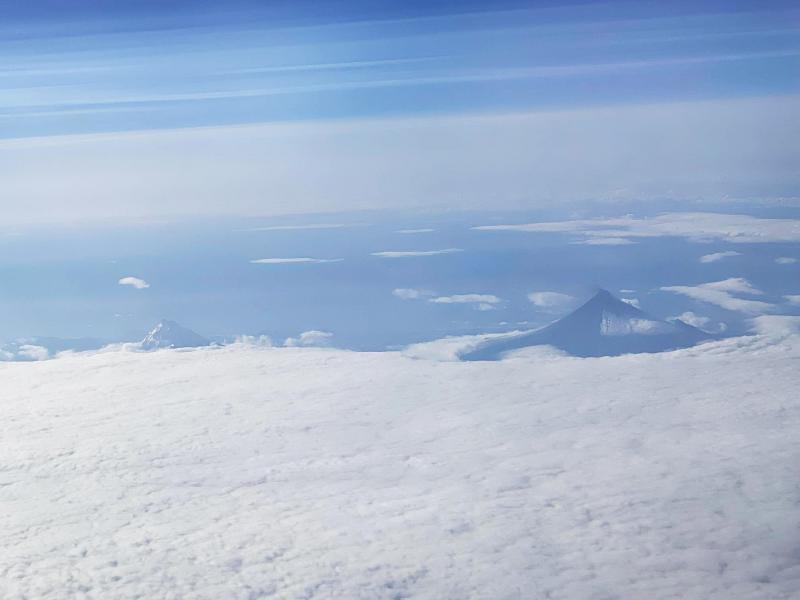 An image of Shishaldin Volcano on Unimak Island, taken from the air on August 15, 2023. The view is from the northeast of the volcano. Most of Unimak Island is cloud-covered in this view, but Shishaldin sticks through the clouds. Dark deposits are visible on its flanks and a small plume of ash is visible from the summit. Isanotski is also visible to the left of Shishaldin.