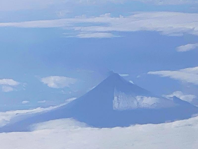 Zoom image of Shishaldin Volcano on Unimak Island, taken from the air on August 15, 2023. The view is from the northeast of the volcano. Much of Unimak Island is cloud-covered in this view, but Shishaldin sticks through the clouds. Dark deposits are visible on its flanks and a small plume of ash is visible from the summit, trailing to the south/southeast.