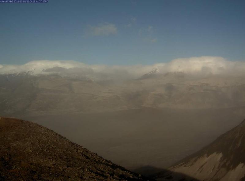 A webcam still from camera KAB2 taken midday on October 2 shows hazy conditions in the Katmai area due to resuspended ash from the 1912 Novarupta eruption. There are two hills in the foreground and more hills in the background-partially snow- and cloud-covered, with a wide flat valley in between. The sky is blue but the whole scene has a fuzzy and gray-ish quality due to the ash.