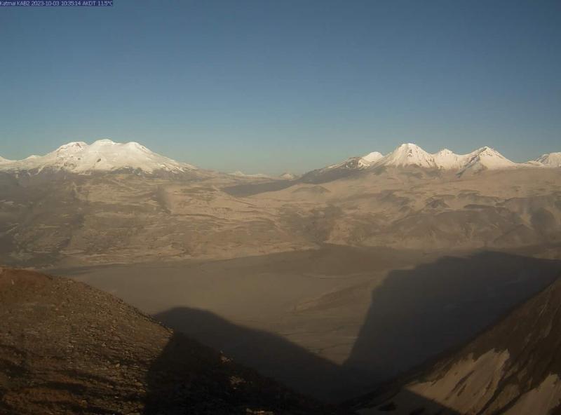 A webcam still from camera KAB2 taken mid-morning on October 3 shows somewhat hazy conditions in the Katmai area due to resuspended ash from the 1912 Novarupta eruption. There are two hills in the foreground and more hills in the background-partially snow- and cloud-covered, with a wide flat valley in between. The sky is blue but the whole scene has a slight fuzzy and gray-ish quality due to the ash.