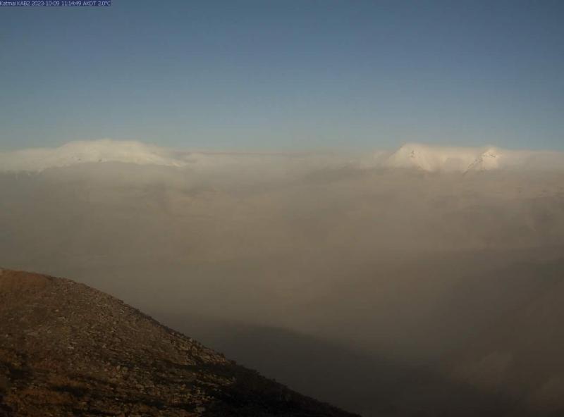 A still from webcam KAB2 in the Katmai area, from 11:14 am AKDT on Monday, October 9, 2023, shows hazy conditions due to resuspended ash. There are two hills in the foreground and more hills in the background that are partially snow- and cloud-covered, with a wide flat valley in between. The sky is blue but the whole scene has a fuzzy and gray-ish quality due to the ash.