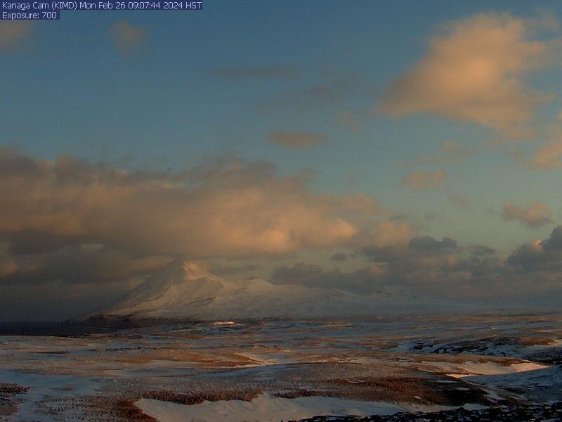Webcam still from Kanaga cam KIMD, taken on February 26 at 9:07 AM local time (10:07 AM AKST). This image shows a sunrise shot of snow-covered Kanaga Volcano in the background, mostly visible amid some low clouds. The foreground is much more flat and dusted with snow between patches of brown winter grass. A small portion of ocean is visible on the left-hand side of the image.