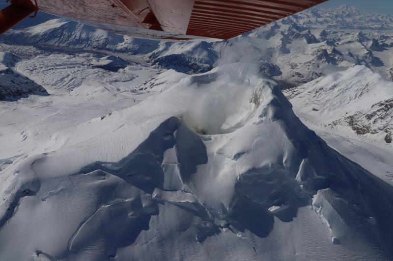 Steaming from the summit crater at Mount Spurr is visible during an overflight on May 14, 2024. The steam source is a fumarole (steam vent) inside of the summit crater, which is snow-free and yellowish, due to sulfur precipitating from the plume. Photograph is taken looking toward the south.