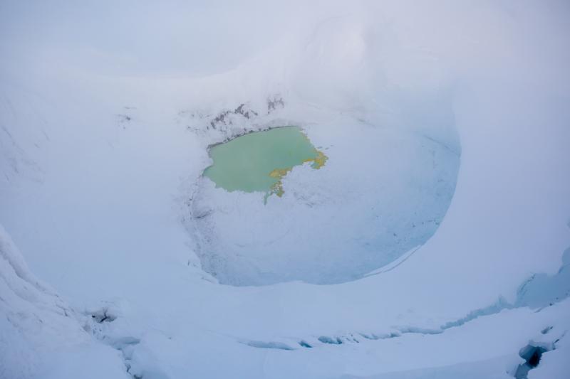 Summit crater of Mount Spurr with a partially ice-covered crater lake on December 18, 2024. Yellow sulfur precipitates are visible floating on the surface. Photo taken part of a photo and gas survey following recent seismic unrest at the volcano.