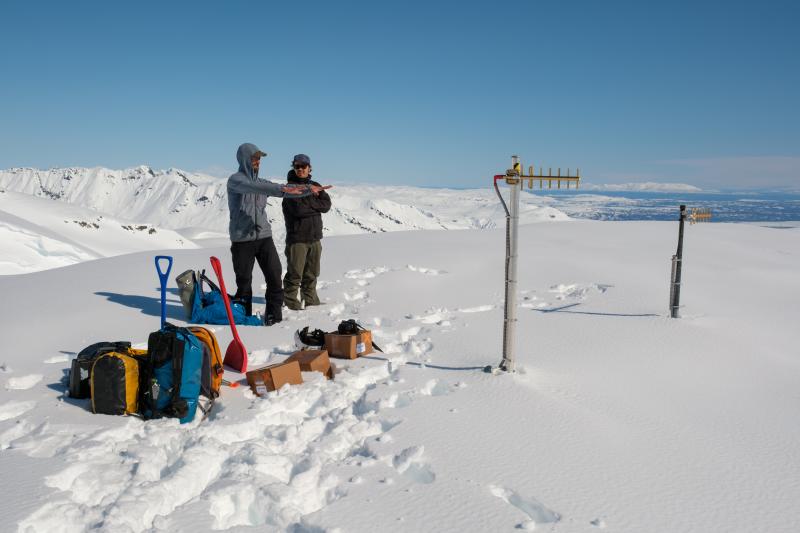 AVO field personnel Dane Ketner and Wyatt Mayo contemplating digging out a buried monitoring hut at site SPCG. The antennae visible above the snow are around 15 feet above the ground surface. The visit purpose was to add batteries and expose solar panels to ensure continued functionality of this station until snow melts in the summer. 