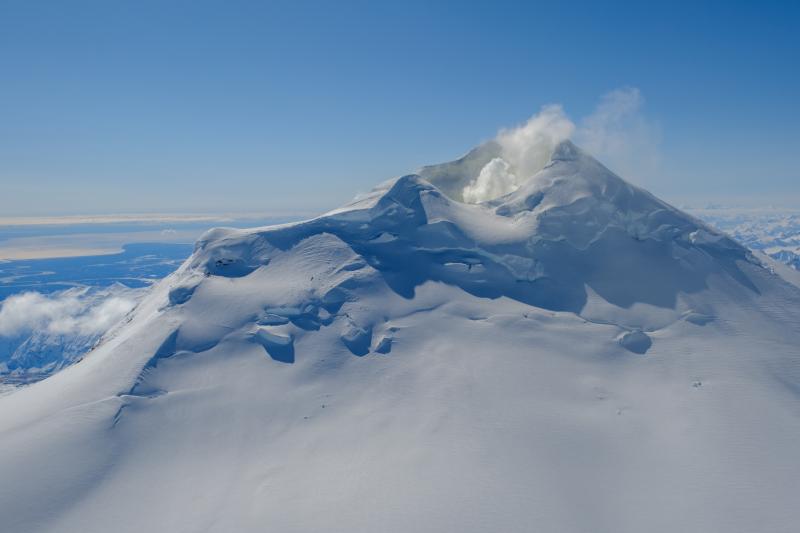 View of the north flank of the Mount Spurr summit cone on April 25 during an AVO gas and photo survey. Steam and gas emissions are visible from the summit crater. Redoubt Volcano is visible on the horizon just to the right of Mount Spurr.