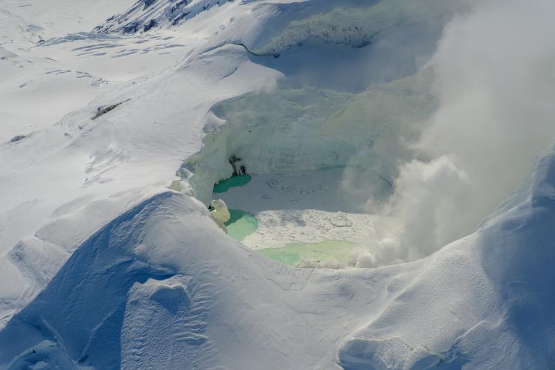 View of the Mount Spurr summit crater lake on an April 25 overflight and gas survey. The water is greenish blue to yellow, due to suspected sulfur precipitates in the water. Floating ice covers the southern half of the lake. View is looking south.