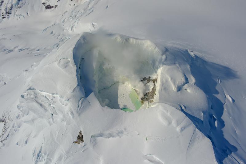 View of the Mount Spurr summit crater lake on an April 25 overflight and gas survey. The water is greenish blue to yellow, due to suspected sulfur precipitates in the water. Floating ice covers the southern half of the lake. The fumarole field on the north shoreline is visibly steaming. 