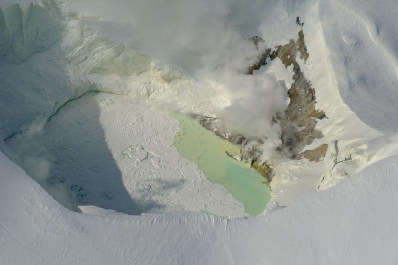 View of the Mount Spurr summit crater lake on an April 25 overflight and gas survey. The water is greenish blue to yellow, due to suspected sulfur precipitates in the water. Floating ice covers the southern half of the lake. The fumarole field on the north shoreline is visibly steaming. 