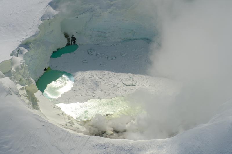 View of the Mount Spurr summit crater lake on an April 25 overflight and gas survey. The water is greenish blue to yellow, due to suspected sulfur precipitates in the water. Floating ice covers the southern half of the lake. The fumarole field on the north shoreline is visibly steaming (bottom of image). 