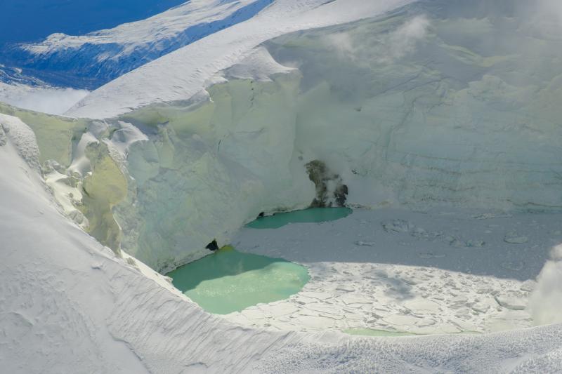 View of the Mount Spurr summit crater lake on an April 25 overflight and gas survey. The water is greenish blue to yellow, due to suspected sulfur precipitates in the water. Floating ice covers the southern half of the lake. The image is looking southeast with two fumarolic areas exposed under recently melted ice visible.