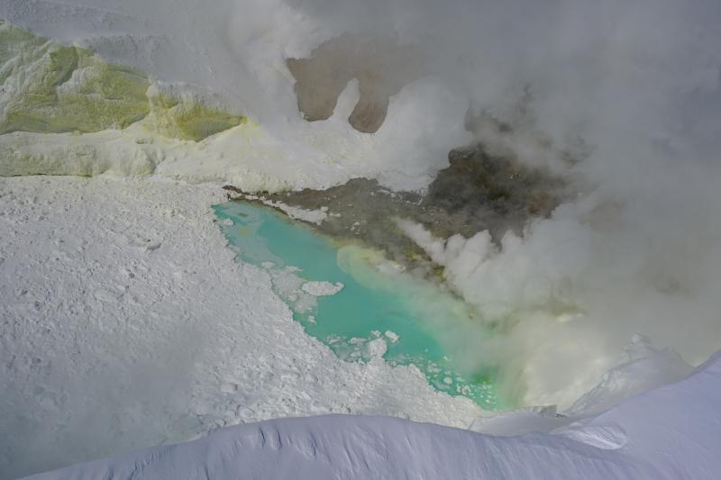 Summit crater of Mount Spurr on May 23, 2025. Gas emissions are visible from steaming fumaroles, primarily on the north shore of the crater lake. The lake continues to have floating ice from ongoing collapse of ice on the inner crater walls, and is a blue-green color. Sulfur staining is visible on the snow inside the crater.
