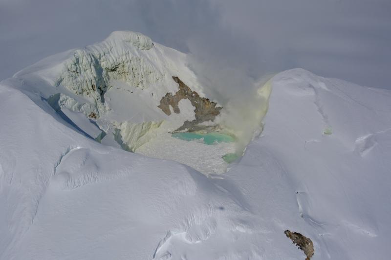 Summit crater of Mount Spurr on May 23, 2025. Gas emissions are visible from steaming fumaroles, primarily on the north shore of the crater lake but also a location on the southeast shore and several smaller fumaroles on the upper flanks. The lake continues to have floating ice from ongoing collapse of ice on the inner crater walls, and is a blue-green color. Sulfur staining is visible on the snow inside the crater.