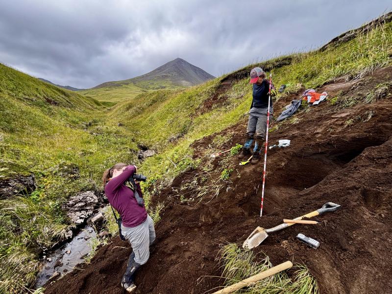 AVO geologists M. Coombs and H. Dietterich photographing the tephra section at station 25AAHD003. Mount Sarichef is in the background. 